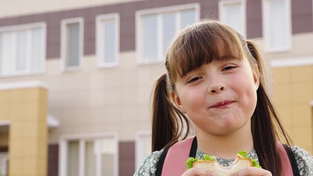 Happy schoolgirl eating sandwich outdoors, Cute child enjoys healthy snack in front of school, Smiling girl with backpack and sandwich, Lunch break happiness for little student, Joyful girl eating