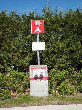 A fire department connection and instructional sign stand in front of green hedges on a sunny day.