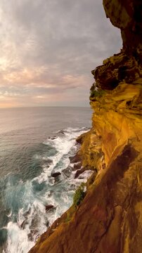Slow-motion wide-angle video of a sunrise over the ocean along the Sydney coastline, with a coastal cliff wall illuminated by soft morning light and calm sea conditions, Australia.