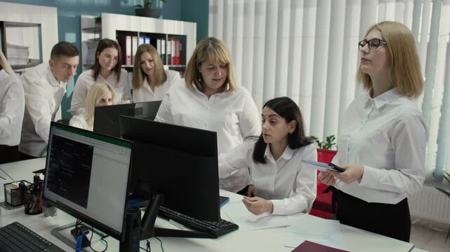 Medium shot, side view, of a team of eight employees, mostly Caucasian, working in an office. All wearing white shirts, analyzing data on monitors. 4K video.