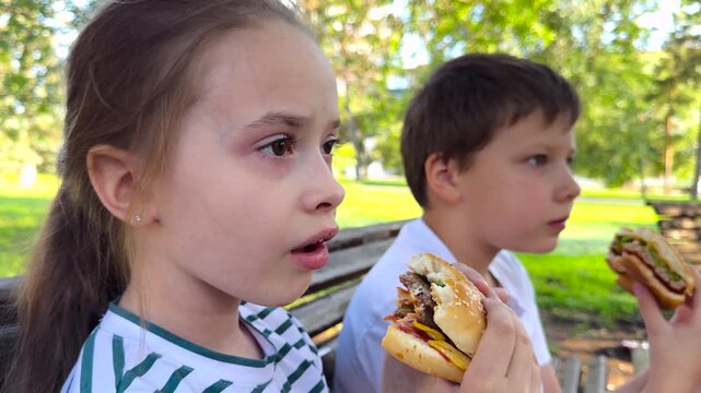 school children eat big hamburger, kids eat burger park, group children friends eat cutlet bun, Kids having hamburgers outdoors, lively lunch in a green park, group gathering on summer day, children