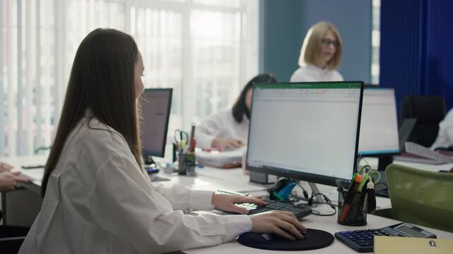 Medium shot, side view, of a Caucasian female employee focused on her keyboard. She is working on a computer in an open office with female colleagues in the bright background. 4K.