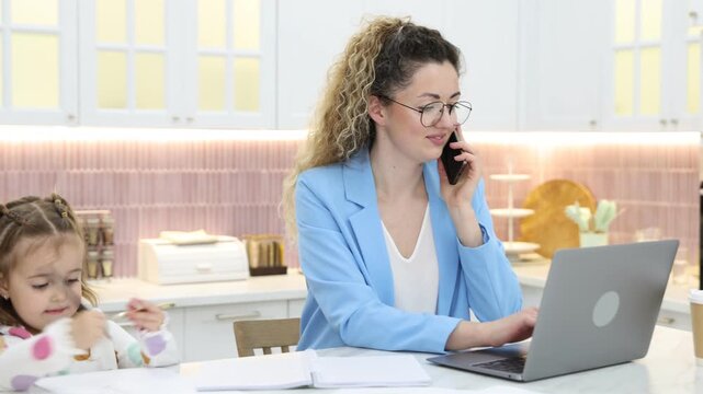 Woman talking on smartphone while working with laptop and her daughter at table in home office, camera moving left