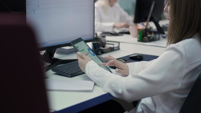 Close-up, side view of a Caucasian female employee using a tablet displaying graphs and a budget. She is sitting at her desk next to her computer monitor. 4K.