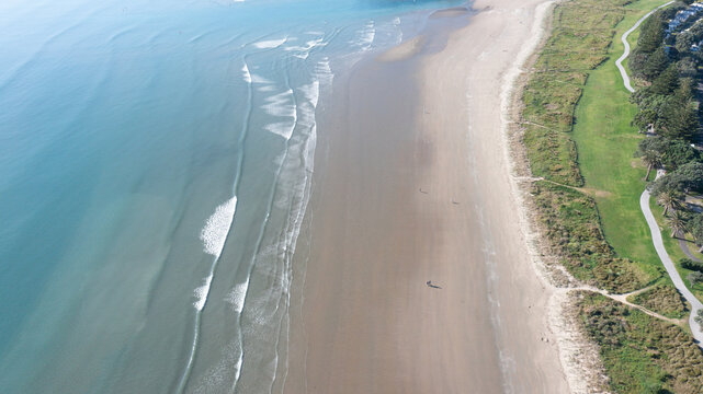 Aerial view of Orewa Beach with gentle waves rolling onto the sandy shore, adjacent to a green coastal park and walking path in Orewa, Auckland, New Zealand.