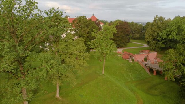 Aerial footage capturing the scenic beauty of the historic Biržai Castle estate in Lithuania. The video showcases the lush green parkland, mature trees, and the distinctive red brick architecture of t