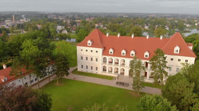 An aerial shot on a bright day shows the reconstructed Biržai Castle manor, a large white two story building with a red tiled roof and an arched arcade facade, surrounded by lush green trees and the t