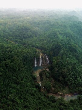 A stunning aerial drone perspective of Curug Cikaso (Cikaso Waterfall) nestled deep within a vast, lush green tropical forest in Sukabumi, West Java, Indonesia