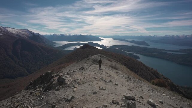 Wide angle shot from behind of a man with a backpack walking down the gravel ridge of Cerro Goye mountain, overlooking snowy peaks and Nahuel Huapi lake in Bariloche, Patagonia.