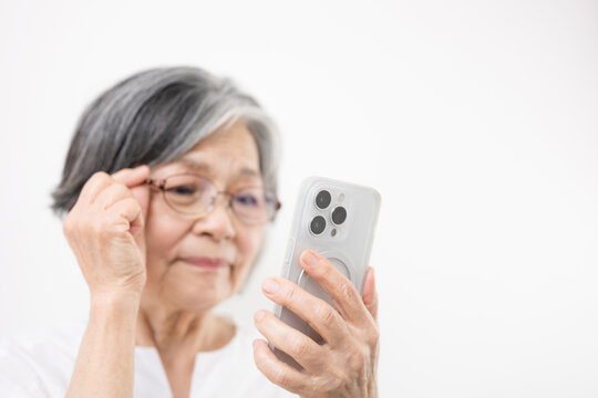 An elderly woman with gray hair, wearing reading glasses and looking at her smartphone against a white background&mdash;an image conveying her struggle to read the text, her suspicion that it might be a sca