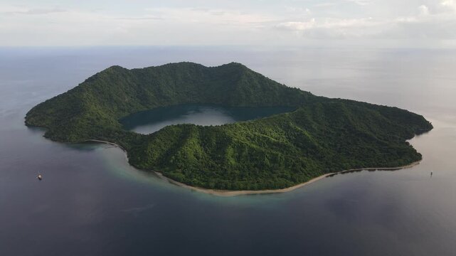 Aerial view of a lush green volcanic island featuring a central crater lake surrounded by calm ocean waters in Indonesia.
