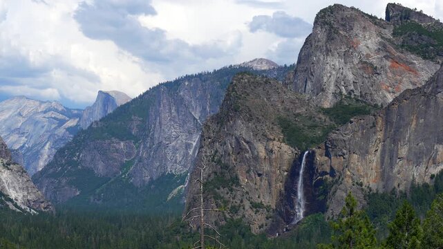 Static shot of Yosemite Valley with a waterfall flowing through granite cliffs and forested landscape in California.