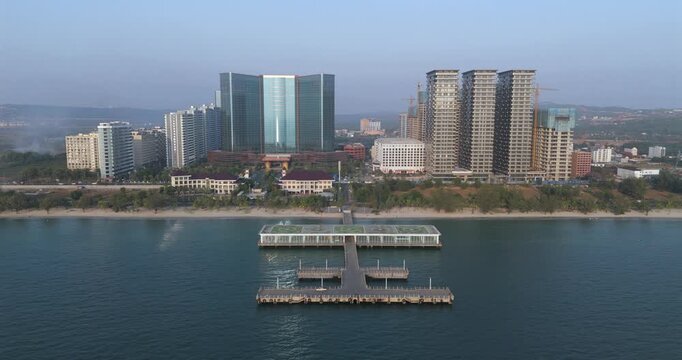 Forward aerial view: a coastal pier with a helipad topped glass terminal extends into the sea, set against the Sihanoukville skyline of high-rise hotels and apartments.