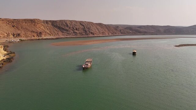 Aerial view of traditional dhow boats floating in the calm waters of a bay surrounded by arid mountains in Sur, Ash Sharqiyah South G, Oman.