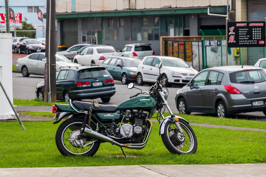 A green Kawasaki motorcycle rests on a grassy verge near auto‑repair businesses in Auckland, surrounded by parked cars and urban street activity
