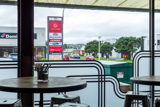 A cosy caf&eacute; interior in Miramar, Wellington looks out toward a busy commercial strip with parked cars, shopfronts, and the Miramar Metro signage listing local businesses
