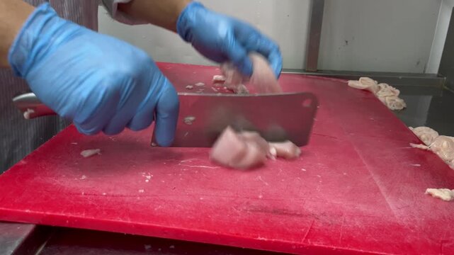 Worker wearing protective gloves chopping meat on a cutting board during food preparation. Raw material processing in a food manufacturing facility following food safety and hygiene practices.