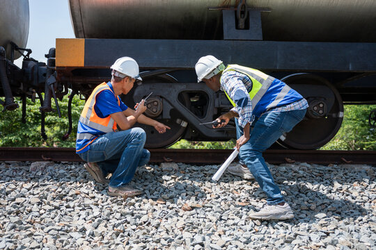 Professional railway engineers checking equipment and logistics infrastructure at train yard, Railway workers inspecting undercarriage and steel wheels of a freight train on tracks