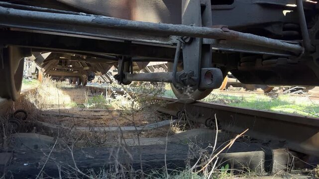 Low view beneath a freight railway wagon showing rusted steel wheels, bogie and suspension above an overgrown disused track. Gritty industrial rail-freight infrastructure