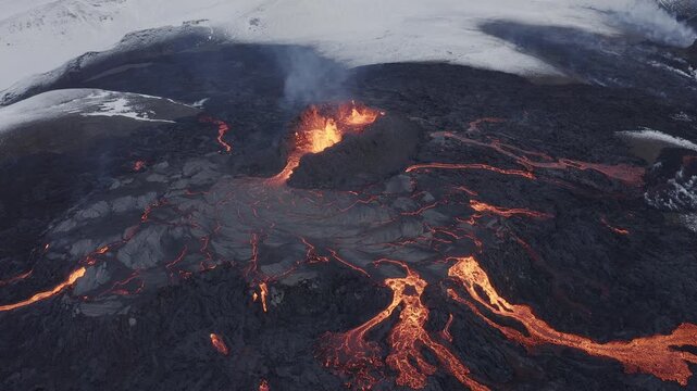 Aerial view of a volcanic eruption with glowing lava flows spreading across dark solidified lava fields and snow-covered terrain in Grimsey, Reykjanesbaer, Iceland.