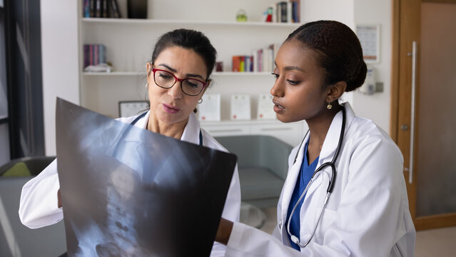 Younger African American doctor asking senior trauma surgeon for advice, teamwork, showing Xray of spine, pointing at image. Two medical professionals discussing scanning exam results. Banner shot
