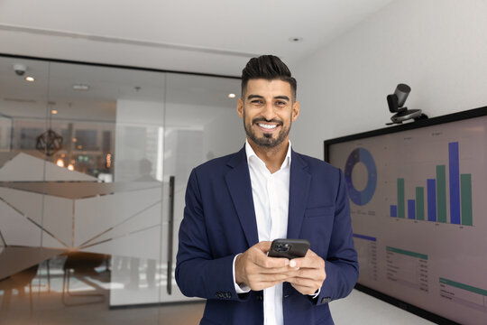 Portrait of smiling Indian businessman in suit looking at camera, standing in alone modern workplace with smartphone, digital screen displaying financial data in background. Tech usage, apps, trading