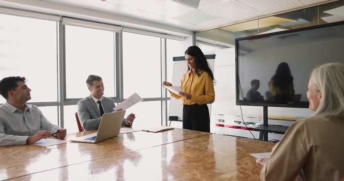 Positive young Indian project manager giving paper reports, marketing research results, presentation slides to coworkers, partners, investors, holding meeting with colleagues