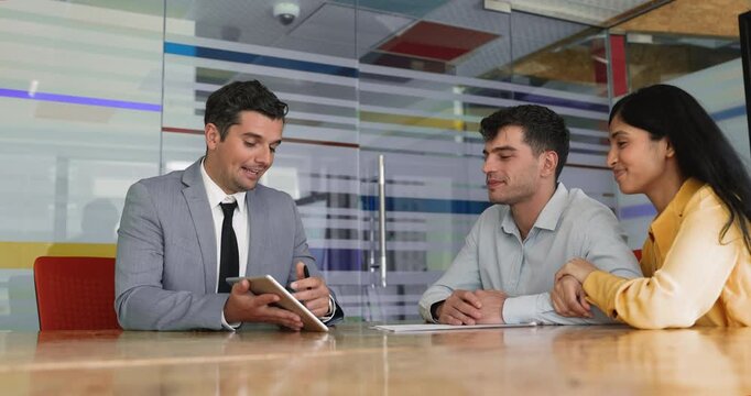 Happy diverse husband and wife visiting lawyer, real estate agent, financial and tax advisor. Friendly male professional showing content on tablet computer to couple of clients