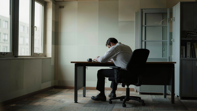 Exhausted businessman slumped alone at desk in dim empty office, workplace burnout and stress concept, editorial business photography with copy space