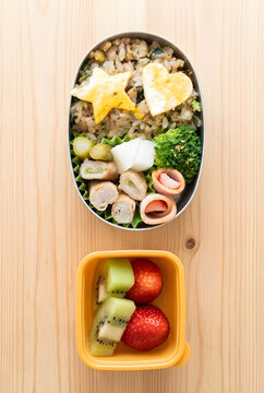 A Japanese homemade character bento lunch box with omurice, heart and star-shaped fried egg, broccoli, and a separate yellow container with kiwi and strawberry fruits on a wooden table, vertical view,