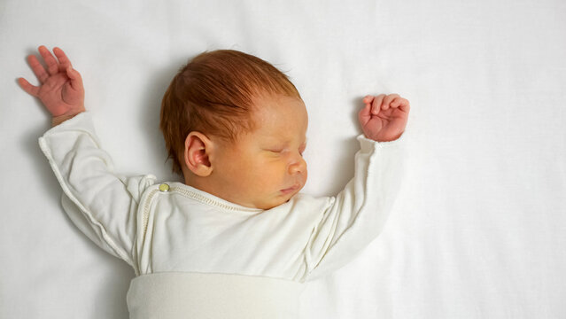 Newborn baby with red hair sleeping profoundly in comfortable white clothes and stretching arms on a clean white sheet, symbolizing innocence, new life, and peaceful dreams