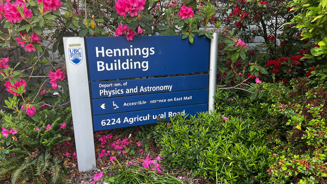 Blue UBC sign for the Hennings Building, home of the Department of Physics and Astronomy at 6224 Agricultural Road, framed by blooming pink rhododendrons on campus.