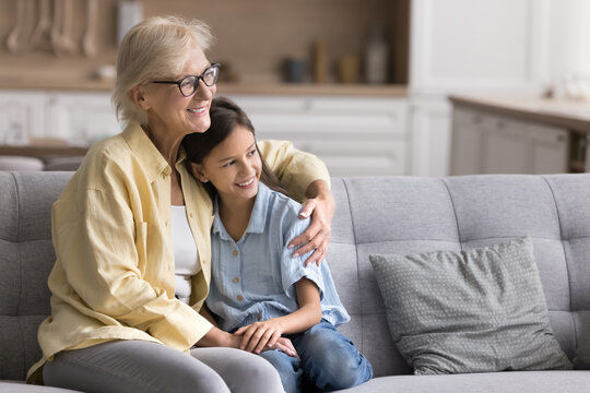 Loving older granny hugging her pretty little granddaughter seated on comfortable sofa looking into distance with positive, optimistic expression, daydreaming, spend priceless time together at home