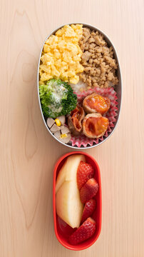 A Japanese homemade bento lunch box with scrambled egg and minced chicken (soboro) on rice, meat-wrapped cherry tomatoes, broccoli, sausage, and a separate red container with rabbit-shaped apple and s
