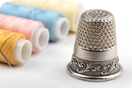 Detailed silver metal thimble featuring intricate decorative floral engravings on the base edge resting on a clean white surface with blurred colorful sewing thread spools arranged neatly in the backg