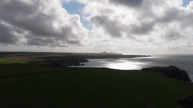Aerial view of the Welsh Coast featuring rugged cliffs and green fields under a dramatic cloudy sky with sunlight reflecting on the sea in St Davids, Wales, United Kingdom.