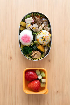 A Japanese homemade bento lunch box with two rice balls (onigiri) - one topped with pink sakura denbu and one with furikake, ginger pork, simmered vegetables, and a separate yellow container with stra