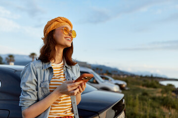 Fototapeta premium Young woman in a denim jacket and striped shirt smiles while looking at her phone beside parked cars by a coastal road.