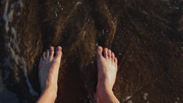 op down view of bare male feet standing on a wet sandy beach as a sea wave washes over them. Crete, Greece