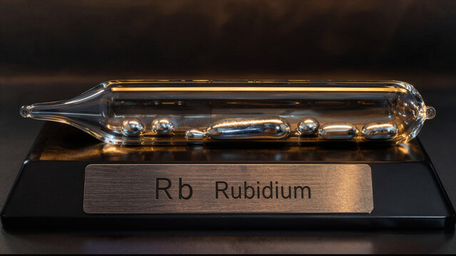 Rubidium is displayed in a glass container with a label showing its symbol and name on a black stand in a laboratory environment