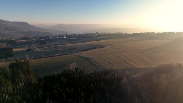 Aerial view of sun-drenched rolling hills and agricultural fields with a small village nestled in the valley during golden hour in Hechingen, Baden-Wurttemberg, Germany.