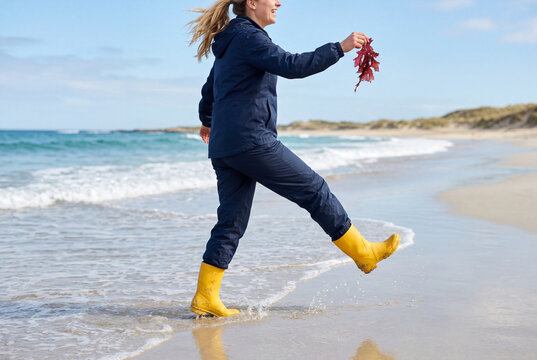 Woman in yellow rain boots kicking water on a sandy beach shore