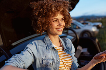 A smiling woman with curly hair in a denim jacket sits in a car, sunlight streams in, reaching toward a handheld device. A relaxed lifestyle scene focused on everyday travel and motion. © SHOTPRIME STUDIO