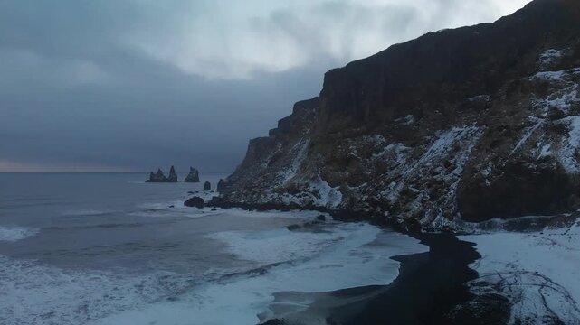 Aerial view of the snow-covered cliffs and black sand beach with sea stacks in the distance under an overcast sky in Vik, Myrdalshreppur, Iceland.