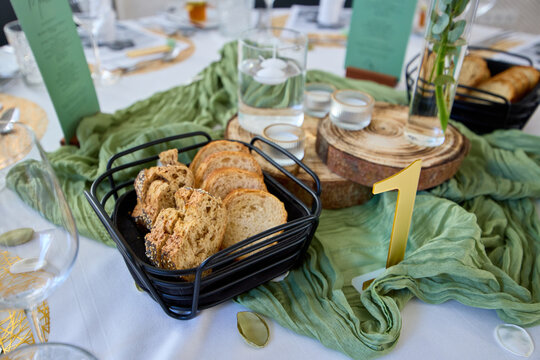 Sliced artisanal bread in a black basket on a wedding table setting with green fabric, wooden coasters, and a gold table number one.