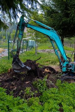 A compact teal excavator grips a large stump by a wire fence, hydraulic arm and hoses visible, soil caked, piles of loam and cut wood ring the pit under conifer branches.