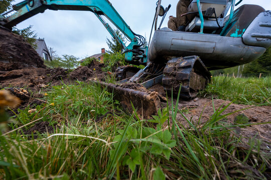 Teal compact excavator carves into wet soil, bucket and steel tracks churn mud near a house roofline. Wide angle close up shows hydraulic arm, worn metal, and crushed grass.