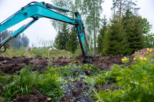 A teal hydraulic excavator digs a narrow muddy trench on a fenced rural plot with conifers and birches, cool overcast daytime, ground level view shows wet soil and water.
