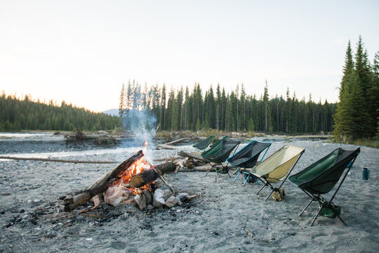 Empty camping chairs around a fire on a riverbank