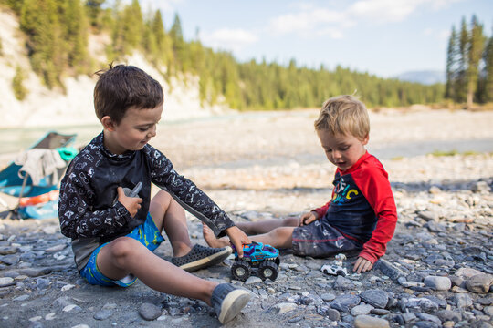 Two young boys play with toy trucks on a riverbank